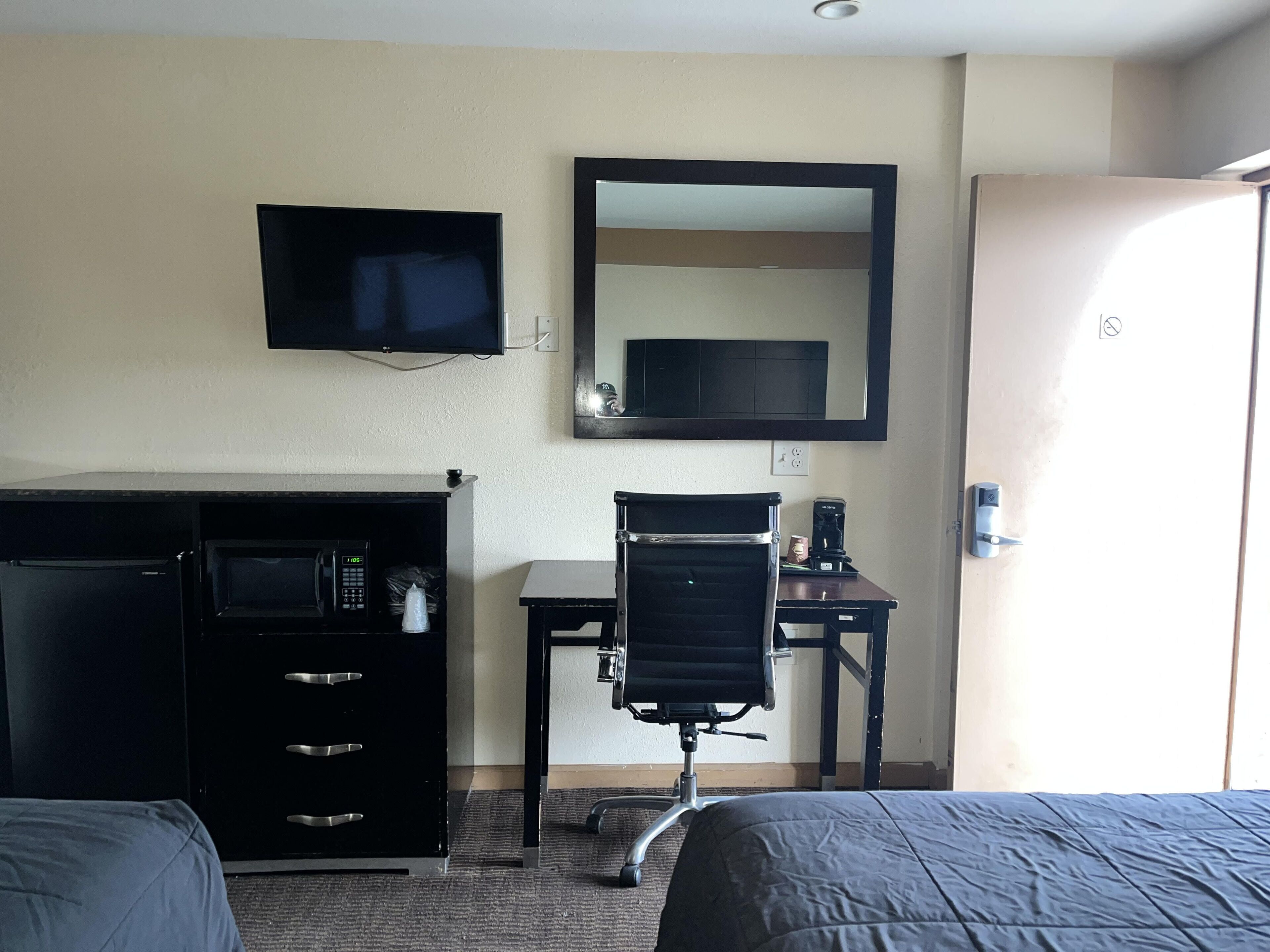 A hotel room with a bed, a desk with a chair, a television, a mirror, and a wardrobe.