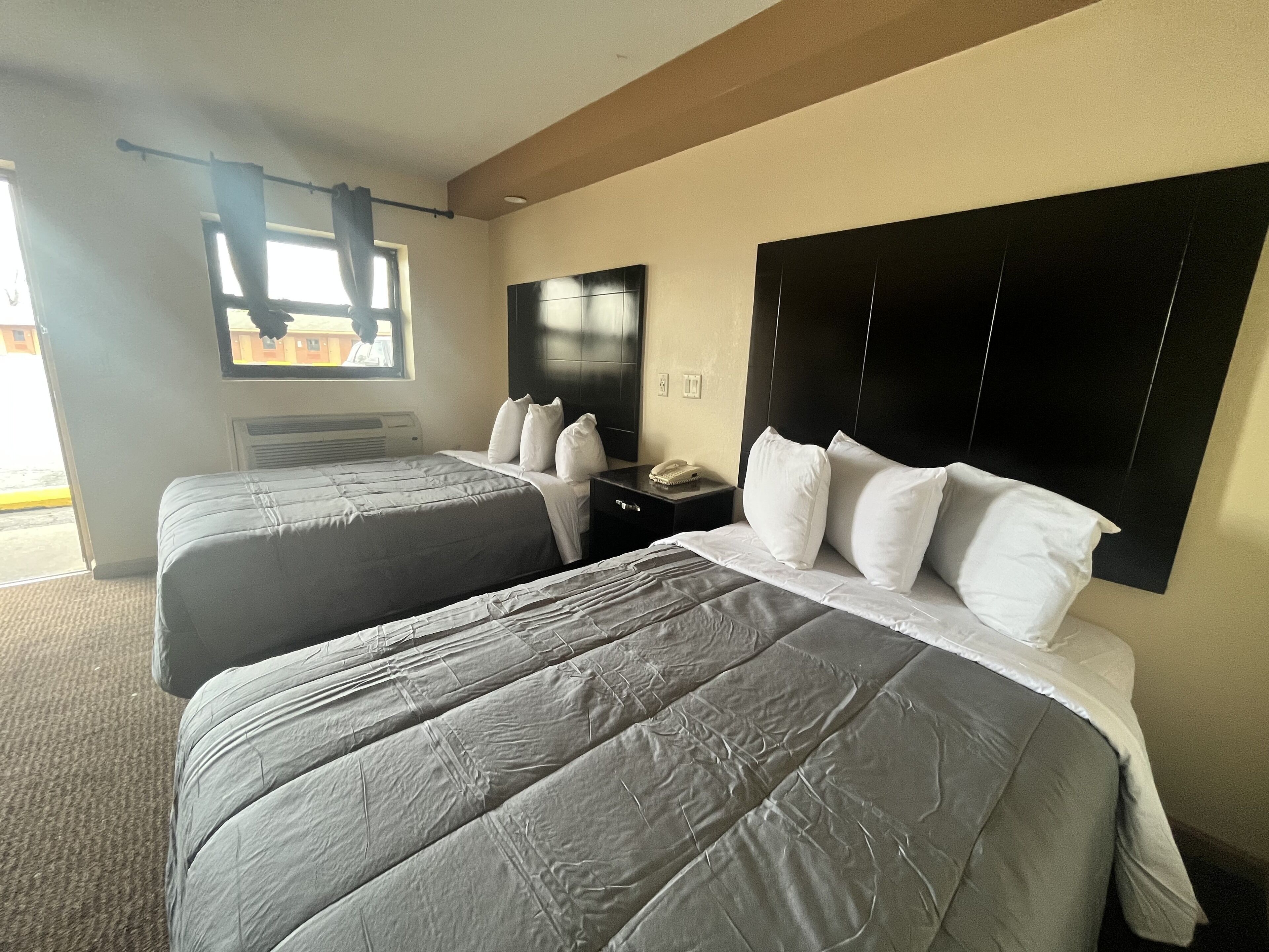 A hotel room with two beds, a large window with curtains, a flat-screen TV, and a bedside table with a phone.