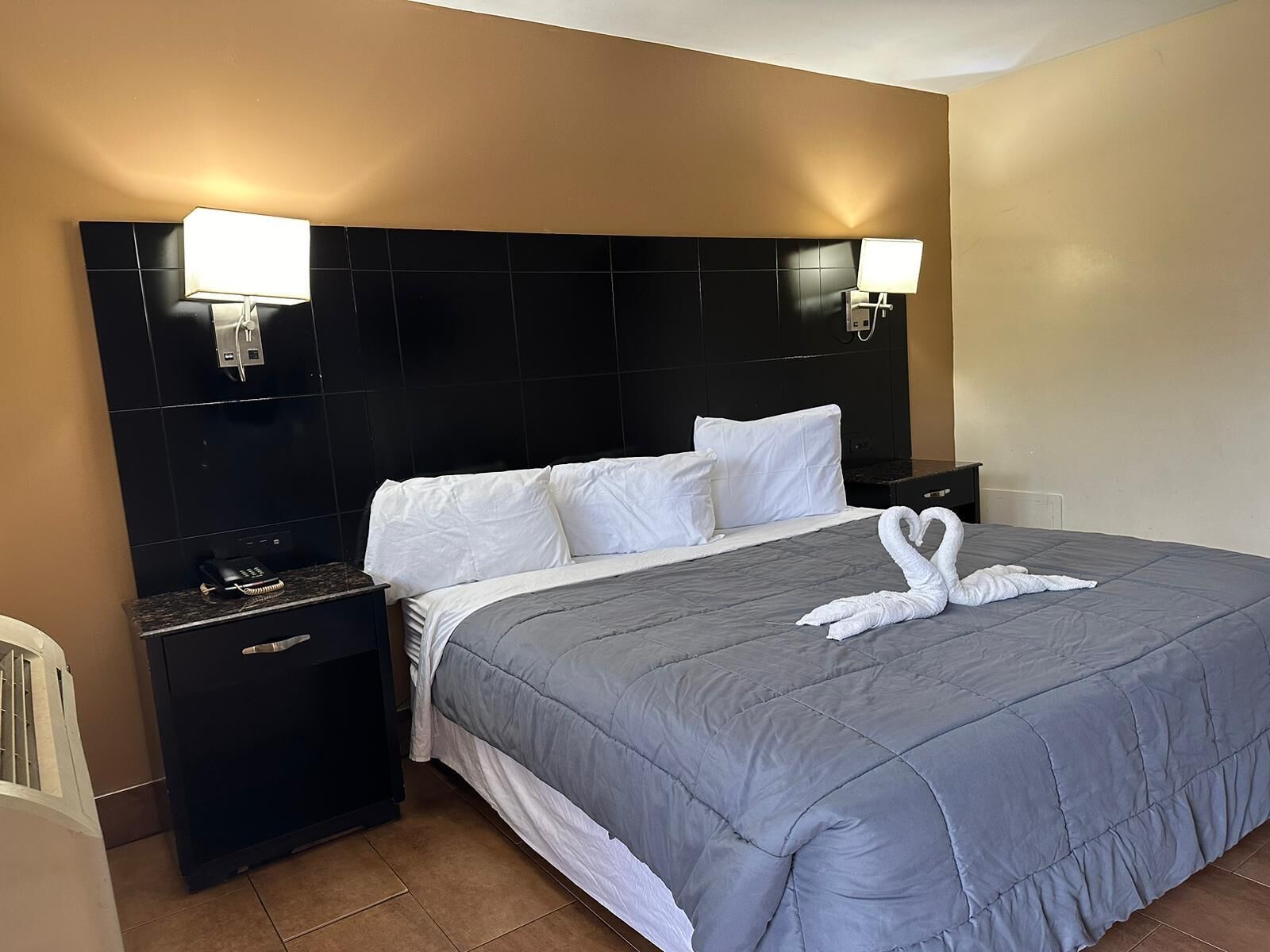 A hotel room with a large bed, two bedside tables, a headboard with wall-mounted lights, and a towel art swan on the bed.