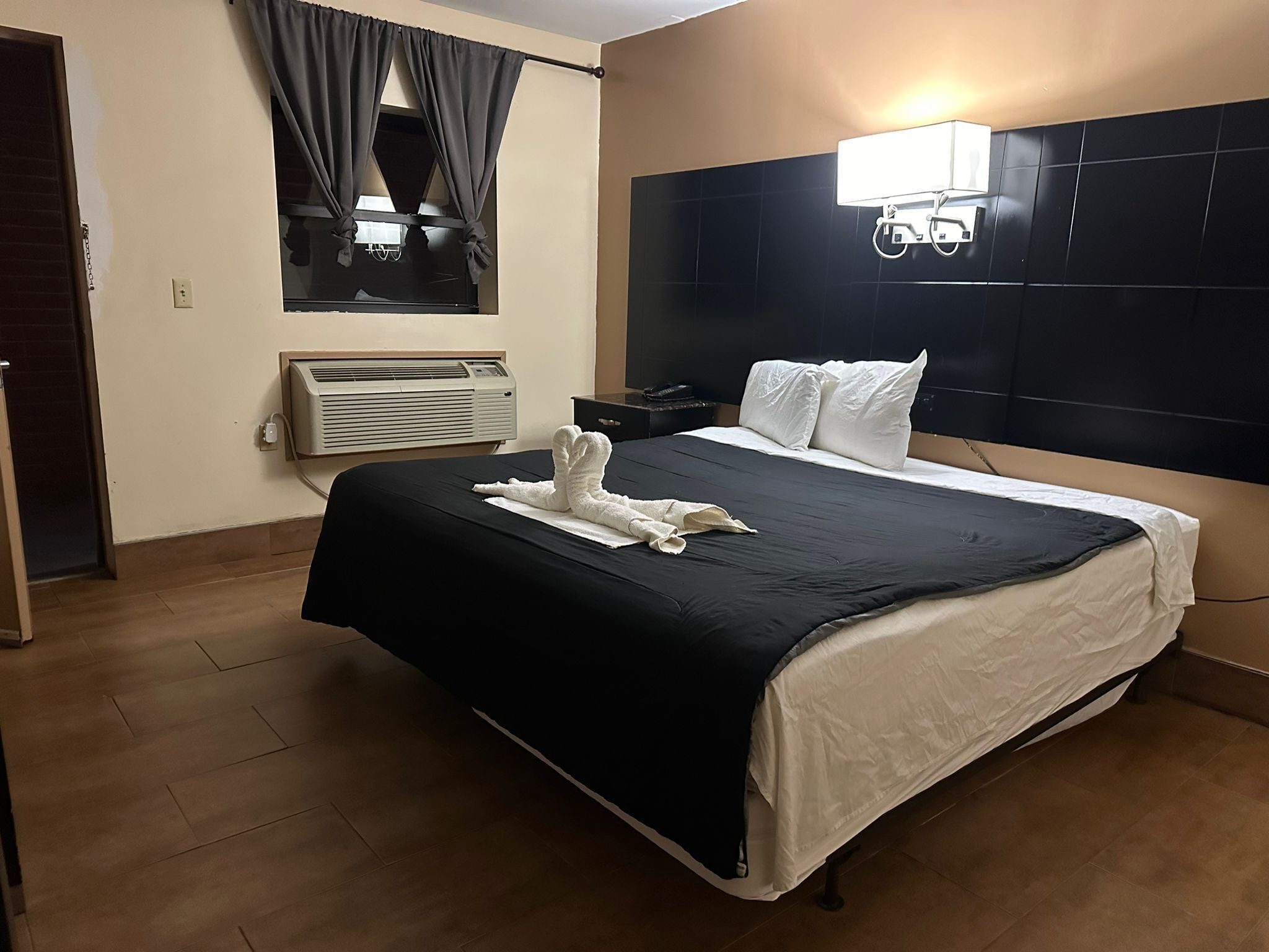 Une chambre d’hôtel avec un grand lit, une tête de lit dotée d’un luminaire, un appareil de climatisation et une fenêtre avec des rideaux.