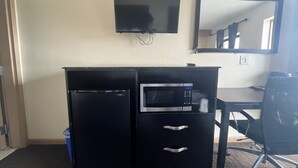 A compact hotel room with a kitchenette, a flat-screen TV mounted on the wall, and a mirror above the desk.