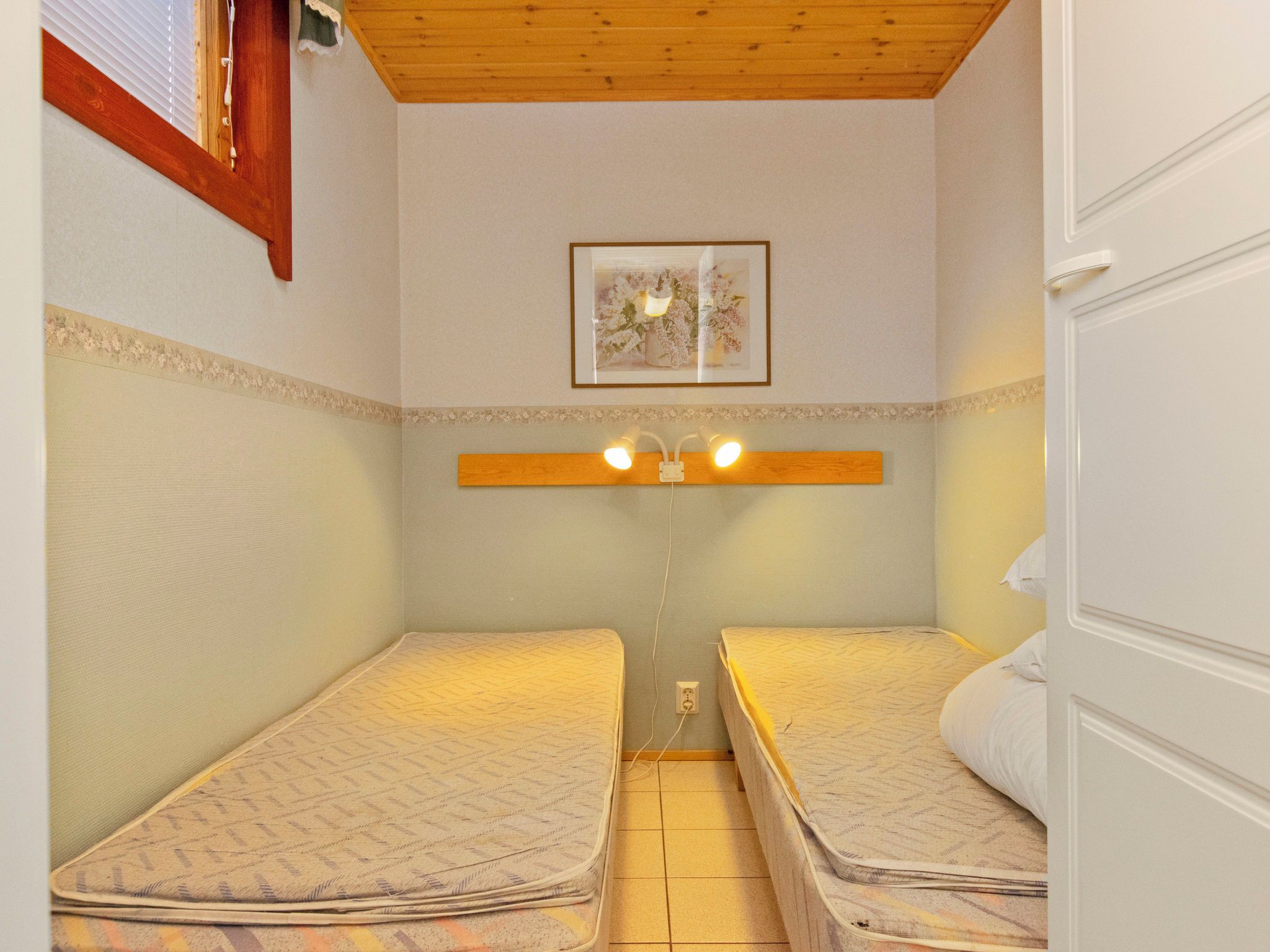 Une petite pièce, de taille modeste, avec deux lits simples, un plafond en bois, un tableau encadré au mur et une porte blanche.