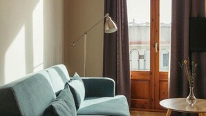Een woonkamer met een bank, een vloerlamp, een klein tafeltje met een vaas, en uitzicht door een raam met gordijnen.