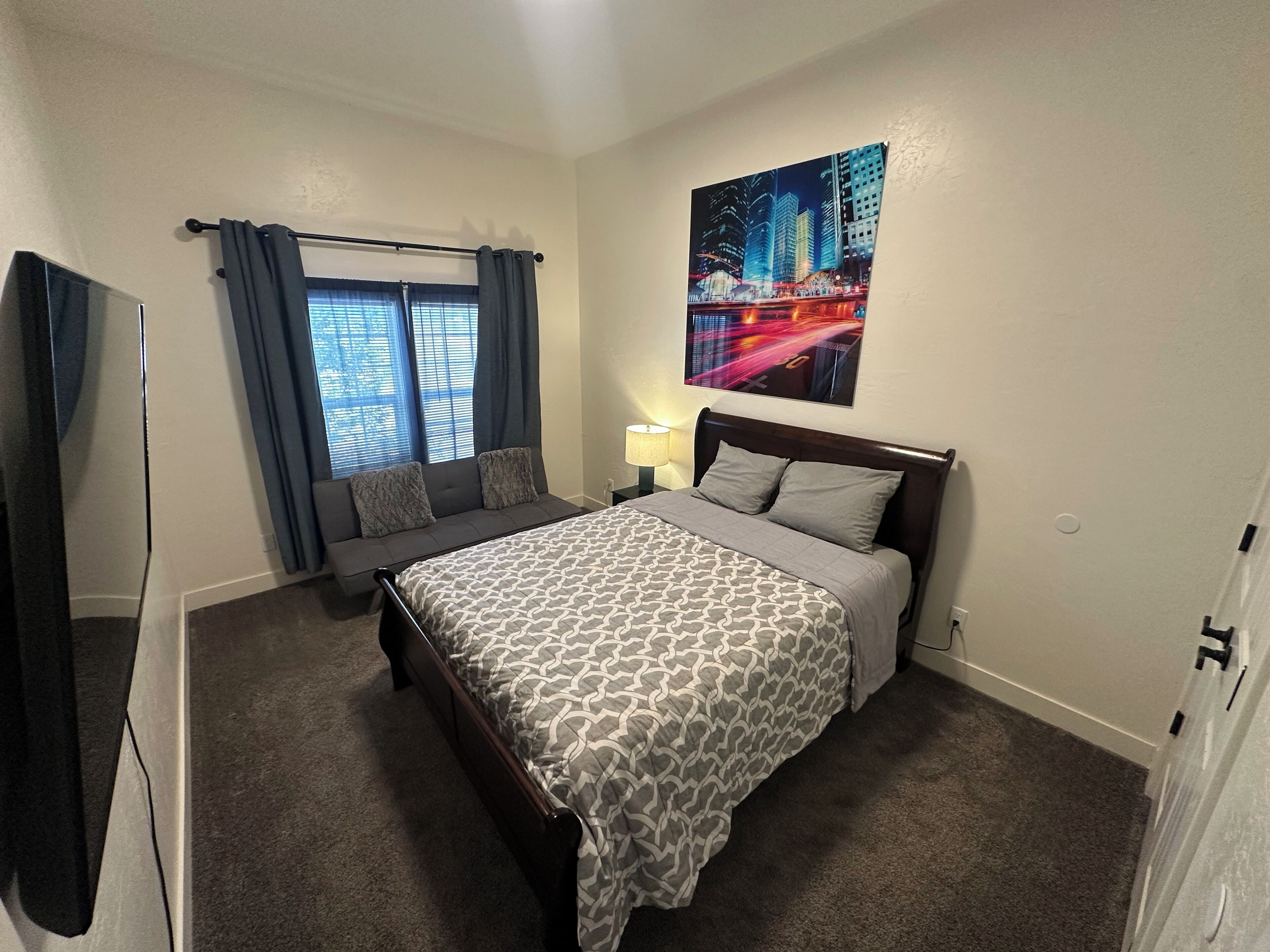 A hotel room with a large bed, a window with curtains, a night cityscape painting, and a lamp.