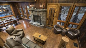 Un intérieur chaleureux avec une cheminée en pierre, un plancher en bois, des canapés en cuir et de grandes fenêtres offrant une vue sur l’extérieur.