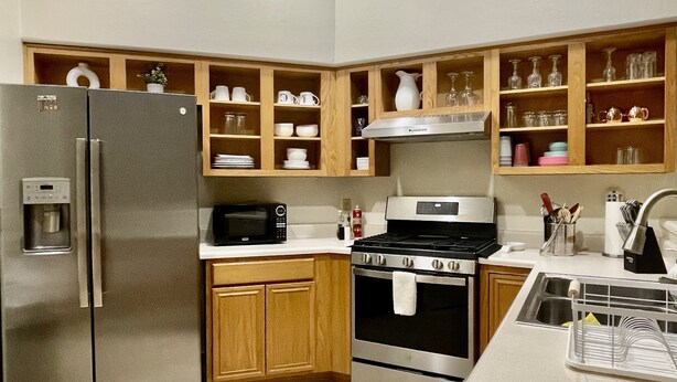 Una cocina moderna con un refrigerador de acero inoxidable, microondas integrado, horno y pileta. Hay armarios y estantes de madera, con diversos utensilios y vajilla.