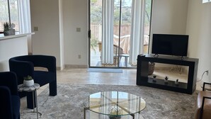 Amplia sala de estar con una mesa de centro de vidrio, sofás oscuros, un televisor sobre un mueble negro y vista a un patio con mesa y sillas cubiertas.