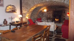Une cheminée en pierre avec un feu de bois, une table en bois et des chaises rouges dans une pièce chaleureuse, agrémentée d’un mur en pierre rustique.