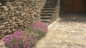 Un escalier en pierre menant à une porte en bois, le tout entouré d’un mur de pierre et de fleurs roses.