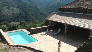 Une piscine en pierre offrant une vue sur une vallée boisée, un patio couvert avec des chaises longues, et une personne se tenant près de la piscine.