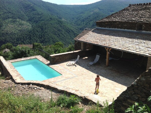 A stone pool with a view of a forested valley, a covered patio with lounge chairs, and a person standing by the pool.