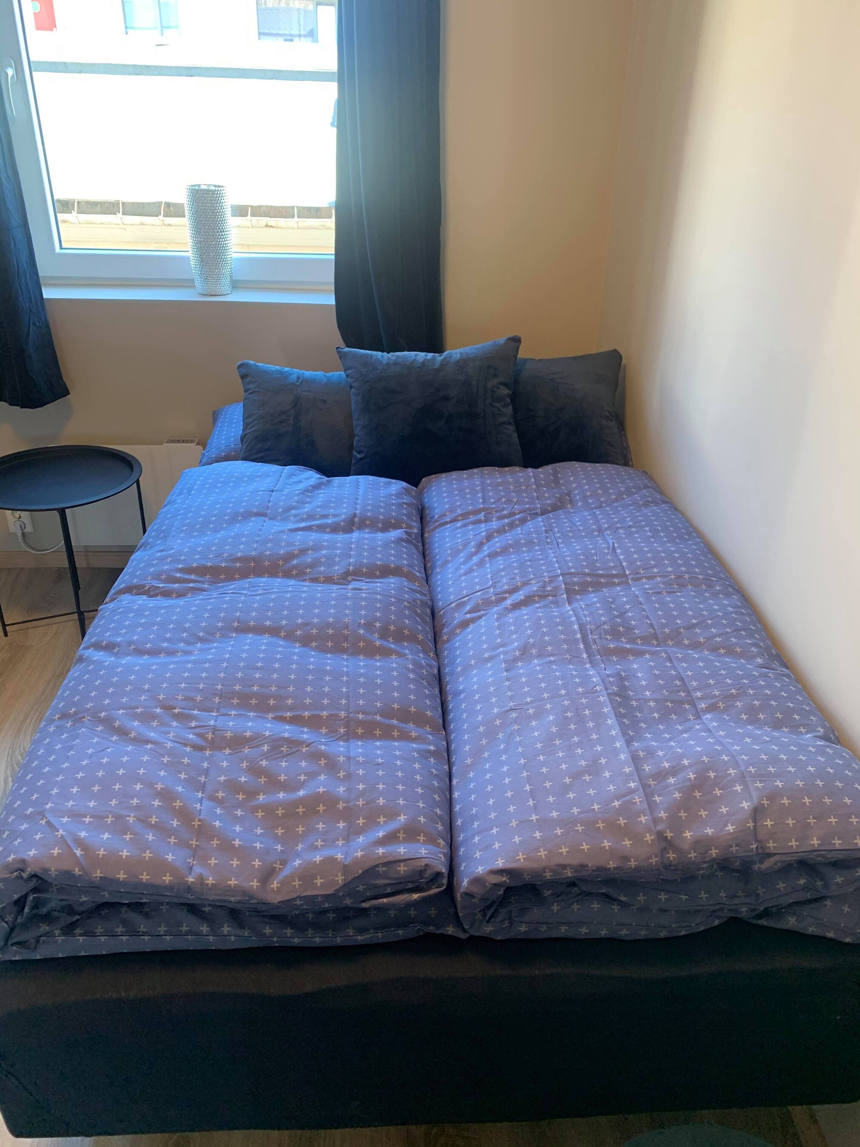 Una cama con colcha estampada, una mesita redonda pequeña y una ventana con cortinas.