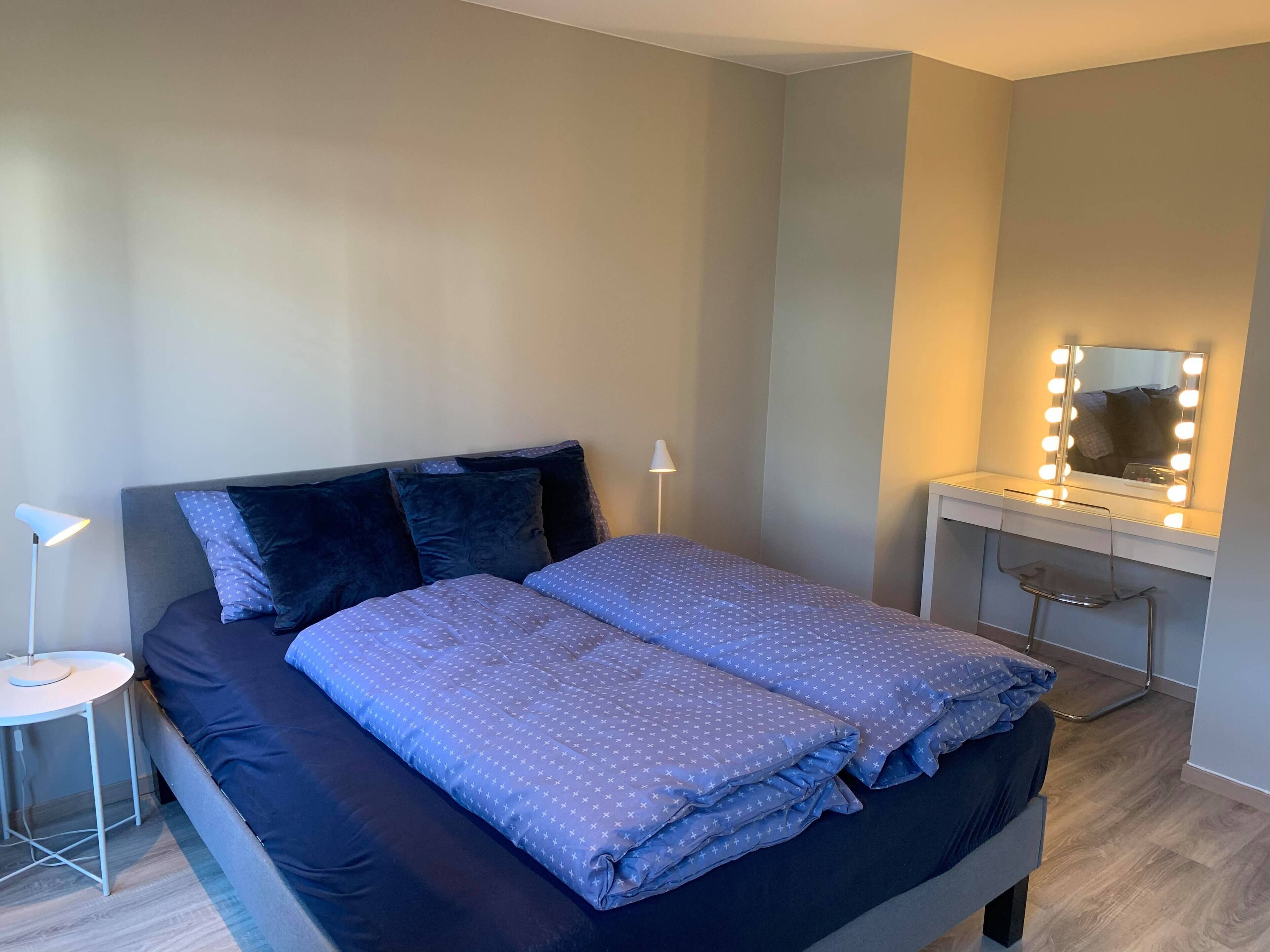 A neatly made bed with a blue patterned duvet, a bedside lamp, and a vanity with a mirror and lights.