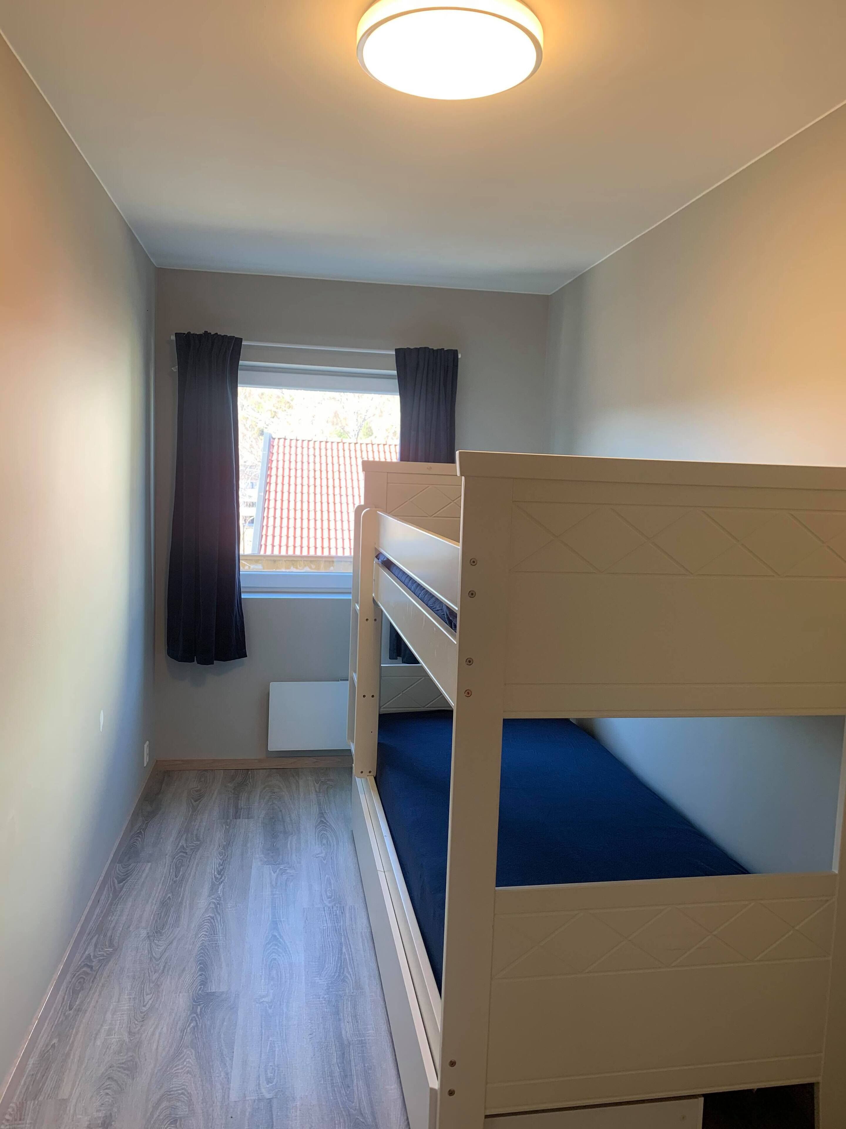 A small, well-lit room with a bunk bed, a window with curtains, and a radiator.