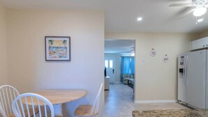 Un comedor con una mesa de madera y sillas blancas, una cocina con un refrigerador blanco, y un pasillo que conduce a otra habitación.