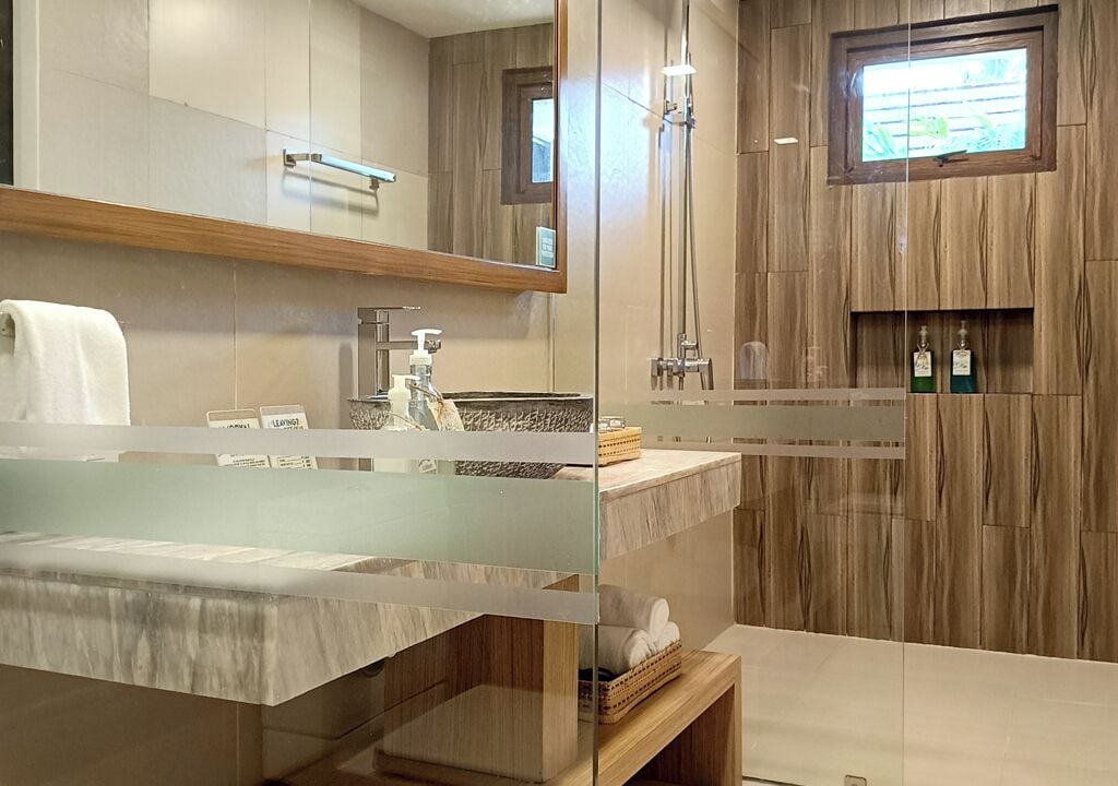 Un baño moderno con mampara de vidrio, tocador de madera, espejo, ducha y ventana.
