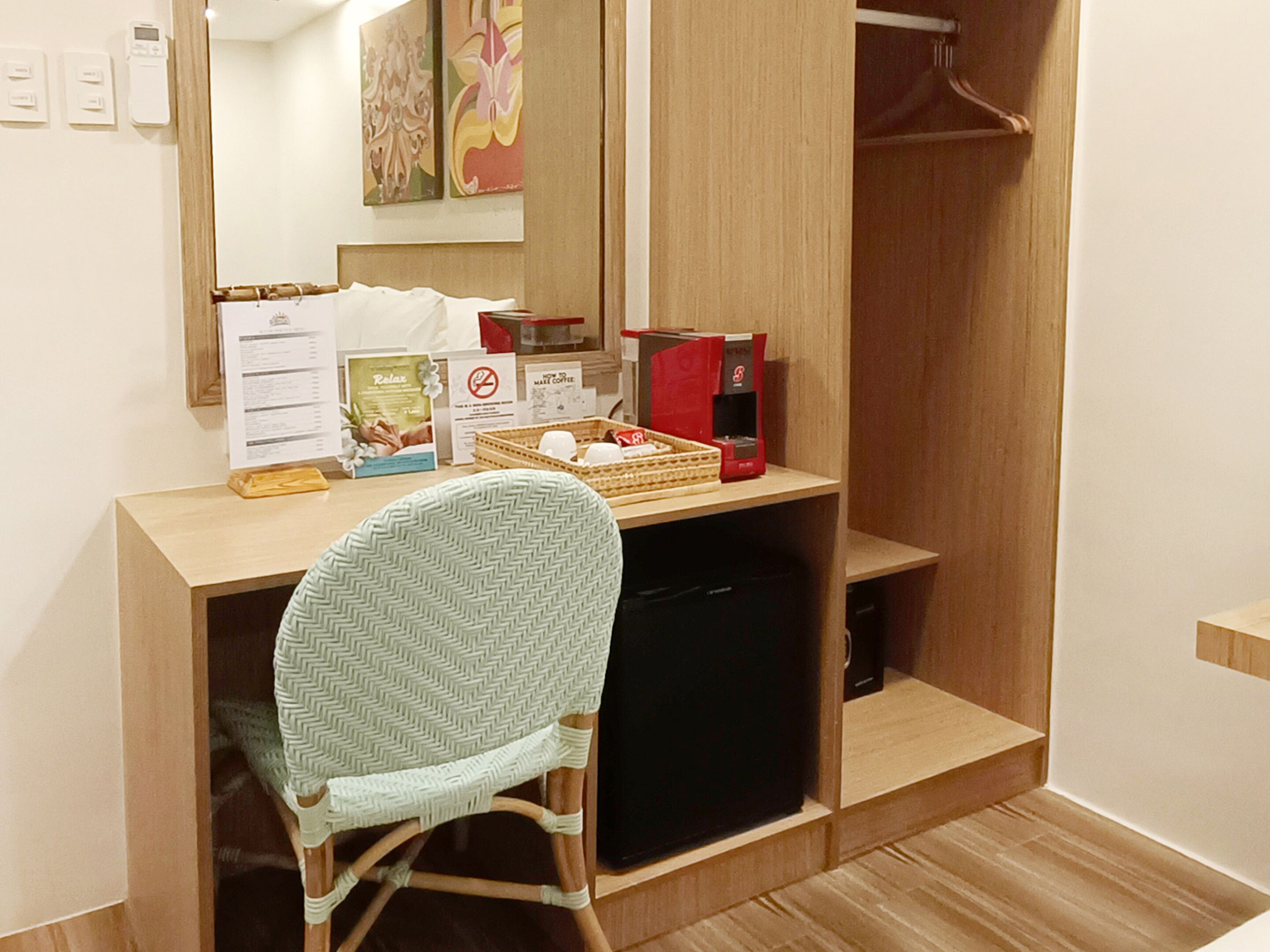 A compact room with a wooden desk, a wicker chair, a coffee machine, and a small black cabinet.