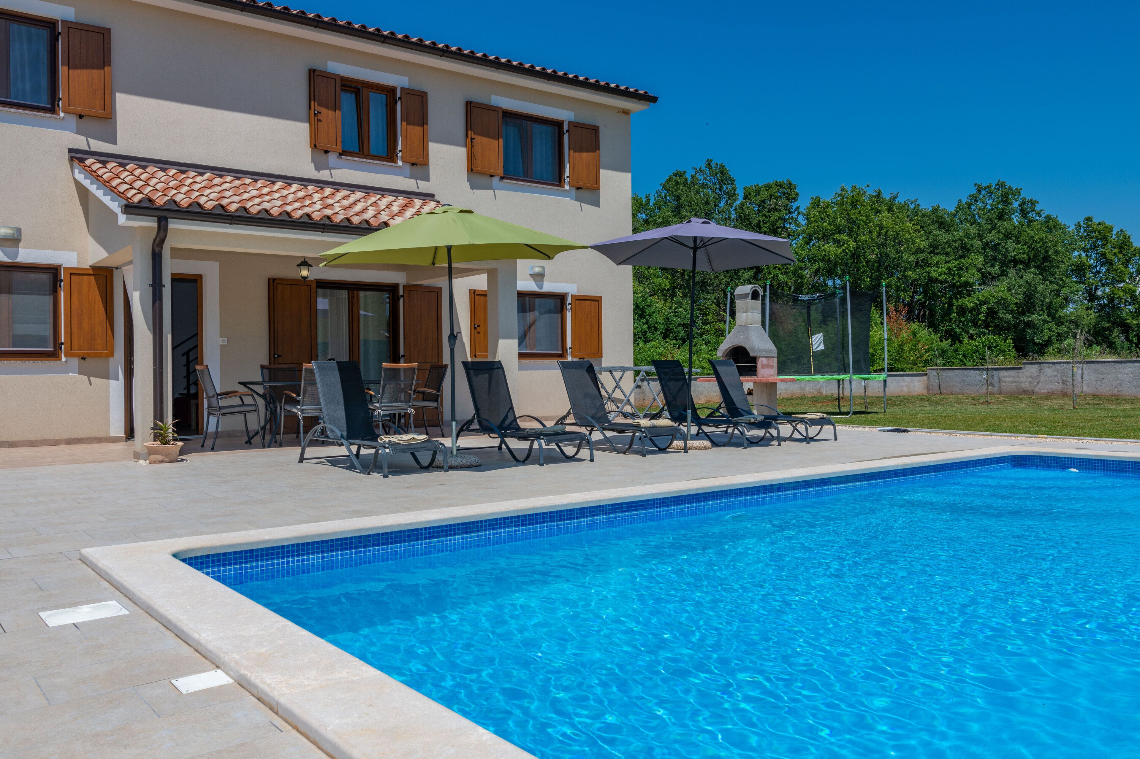 A swimming pool with lounge chairs and umbrellas, a covered patio area, and a building with multiple windows and doors.