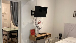 Et hotelværelse med en seng, et skrivebord med stol, et fjernsyn ophængt på væggen og et lille bord med en vase samt et skilt.
