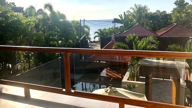 Un balcón con barandilla de vidrio da a una zona de piscina, rodeada de exuberante vegetación y vistas al mar.