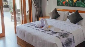 Habitación de hotel con una cama grande, un cuadro de flores tropicales, cabecera de madera y vista a un jardín por una puerta corredera.