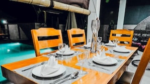 Mesa de comedor para cuatro personas con sillas, piscina al fondo y techo de bambú.