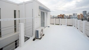 Une terrasse sur le toit, dotée d’une balustrade blanche, d’un appareil de climatisation et offrant une vue sur le paysage urbain en arrière-plan.