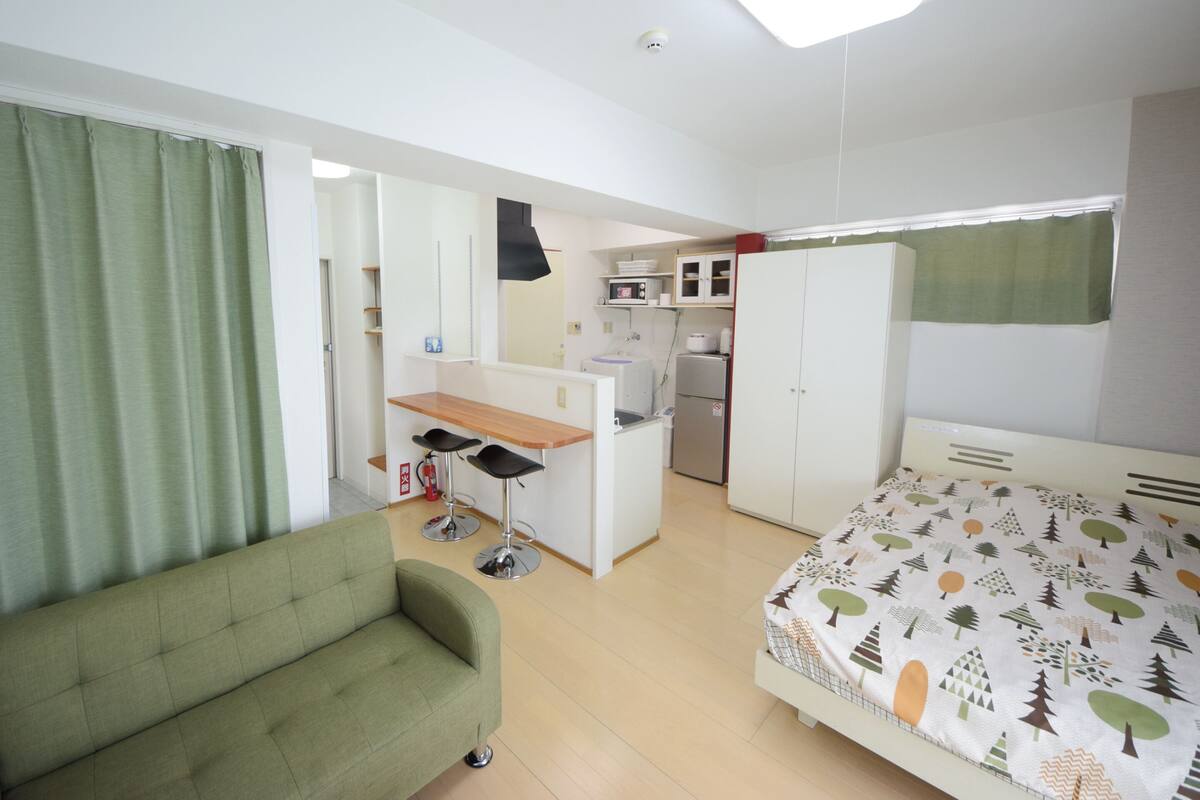 A compact room with a bed, a green sofa, a kitchenette, and a small desk.