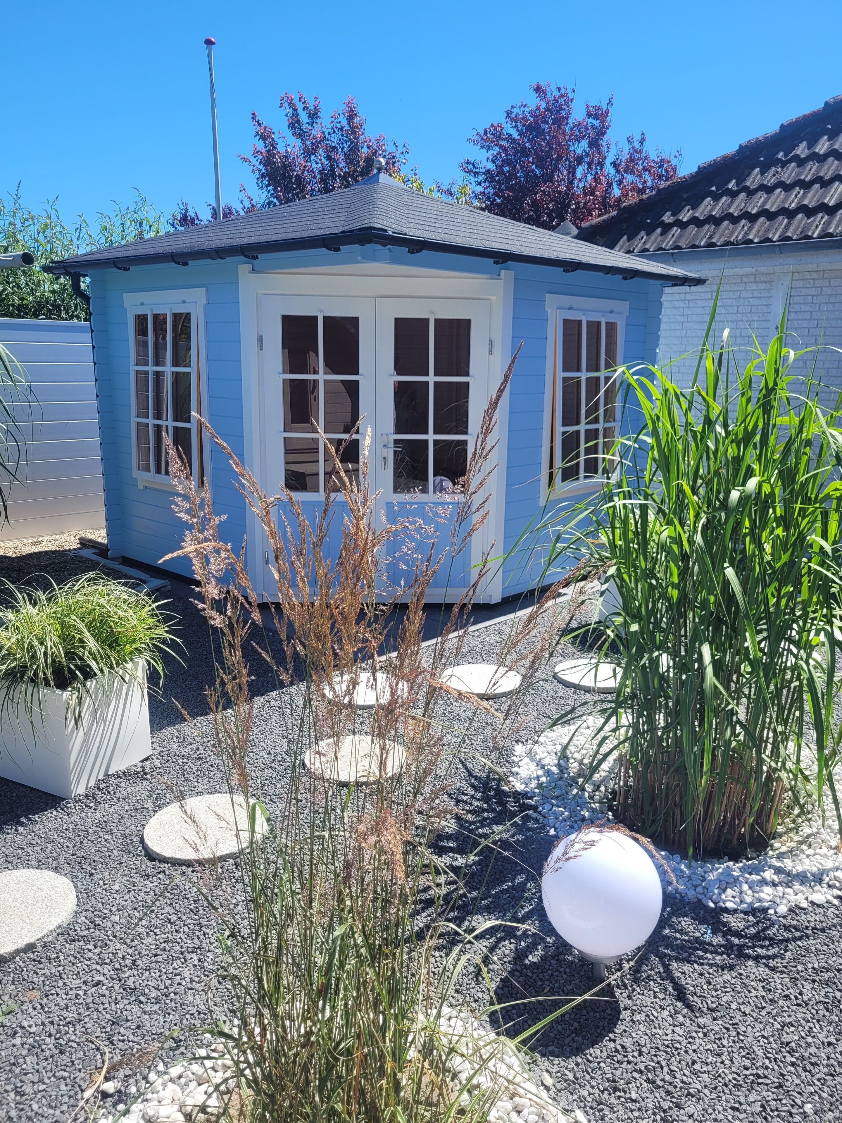 Een klein blauw huis met veranda, omgeven door grind en planten.