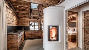A log cabin interior with a central fireplace, wooden walls, a kitchen area, and a sleeping area.