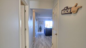 Un pasillo con un letrero de madera que dice "TO BEACH" en la pared, que conduce a una habitación con ventana y planta.