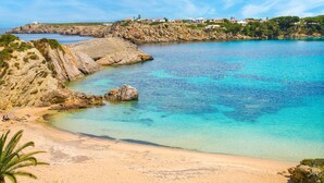 A coastal beach with clear turquoise waters, rocky cliffs, and a distant view of houses on a hill. - Apartment "White Sands 306" on the Beach with Sea View, Pool (Arenal d'en Castell)