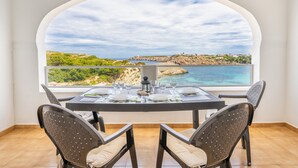 A dining area with a table set for four, a view of the sea and cliffs, and a large arched window. - Apartment "White Sands 306" on the Beach with Sea View, Pool (Arenal d'en Castell)