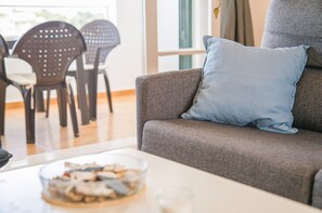 A living room with a grey sofa, a blue pillow, a glass bowl with snacks, and a dining area with black chairs.