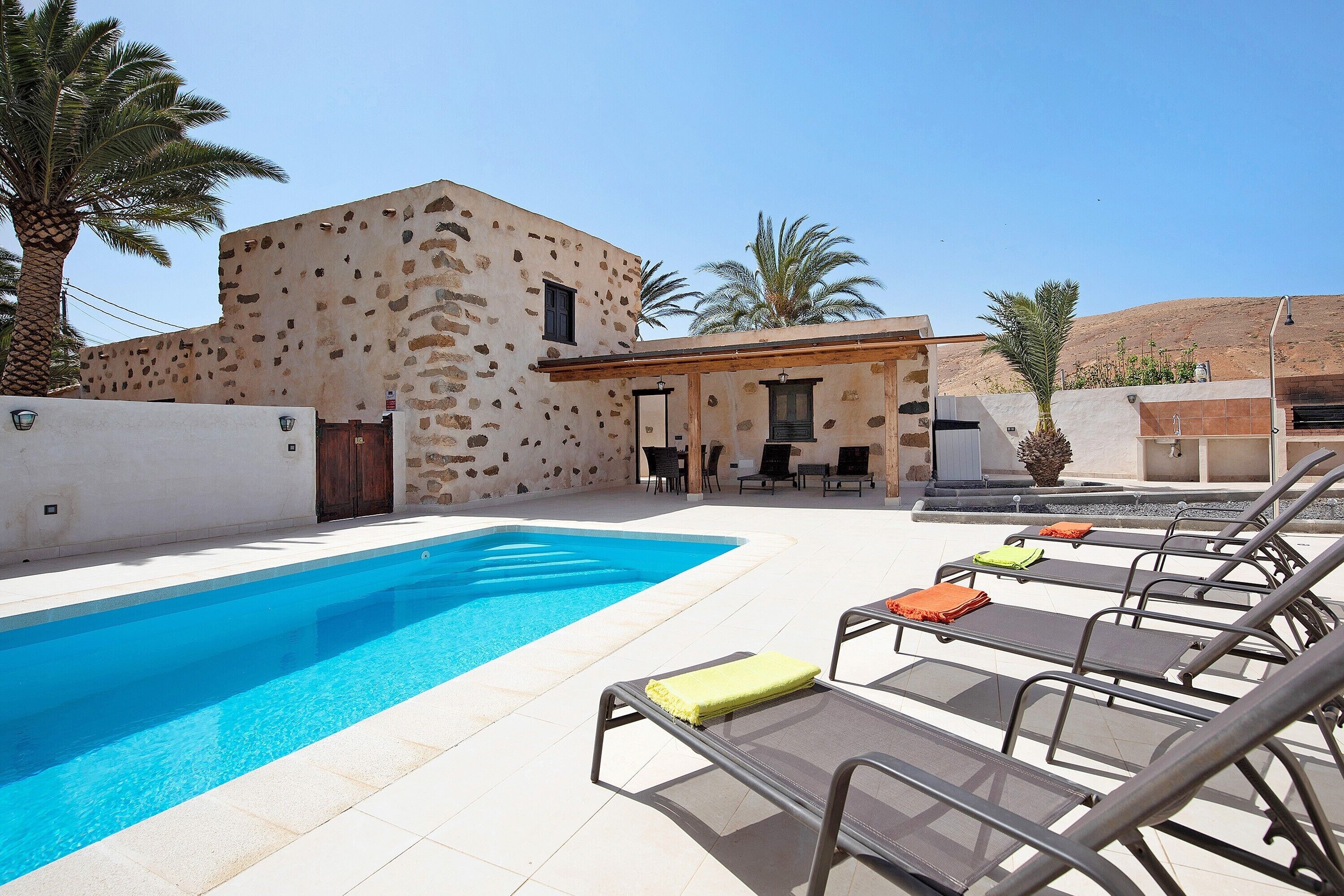 A swimming pool with lounge chairs, a building with stone walls, and palm trees.