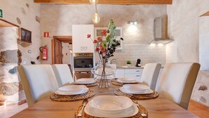 A dining area with a wooden table set for four, surrounded by white chairs, in a room with stone walls and a kitchen in the background. - "Casa Rural Andresito" with Mountain Views, Pool, Wi-Fi, and Terrace (Pájara)