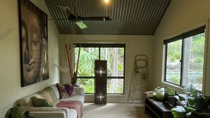 A living room with a high ceiling, a sofa, a coffee table, a large window, and a Buddha painting. - 2 x queen bedrooms, 2 x ensuite's, 1 guest lounge, outdoor bbq kitchen and pool. (Currumbin Valley)