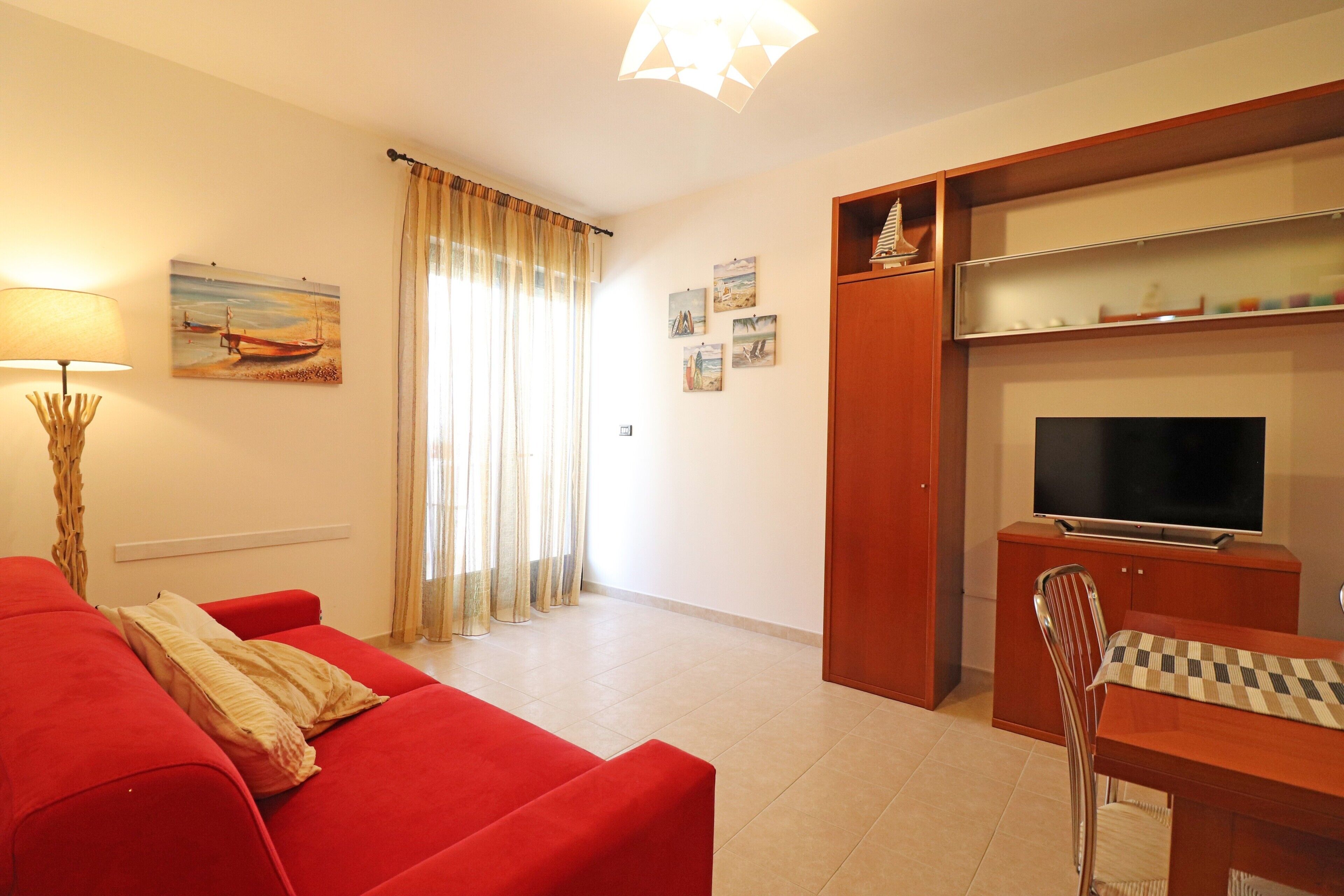 Una sala de estar con un sofá rojo, televisión, un tablero de ajedrez y una ventana con cortinas.