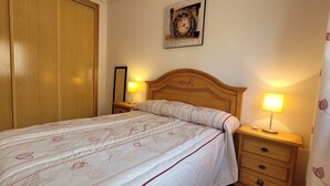 Ein ordentlich bezogenes Bett mit einem gemusterten Überwurf, einem Holzkopfende, Nachttischlampen, einem Nachttisch mit Schublade und einem Holzschrank.