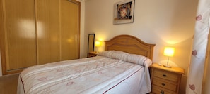 A neatly made bed with a patterned bedspread, a wooden headboard, bedside lamps, a nightstand with a drawer, and a wooden wardrobe.