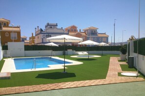 Une piscine avec un plongeoir, entourée de gazon synthétique vert et de parasols blancs.