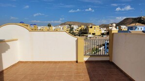 Eine Terrasse mit Fliesenbelag, einem Metallgitter und Blick auf eine Wohngegend mit Gebäuden und einer Hügelkulisse im Hintergrund.