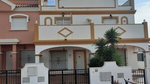 Ein mehrgeschossiges Wohngebäude mit einer symmetrischen Fassade, das einen zentralen Eingang mit befestigter Auffahrt, einen Balkon im oberen Geschoss und eine kleine Gartenanlage mit einer Palme zeigt.