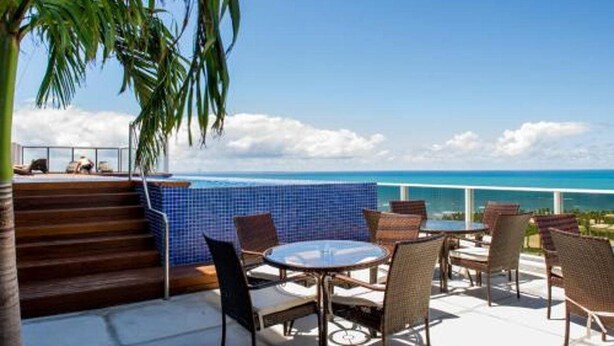 A terrace with a view of the ocean, featuring a round table and wicker chairs, and a blue-tiled wall. - Tryp Pernambuco (Jaboatao dos Guararapes)