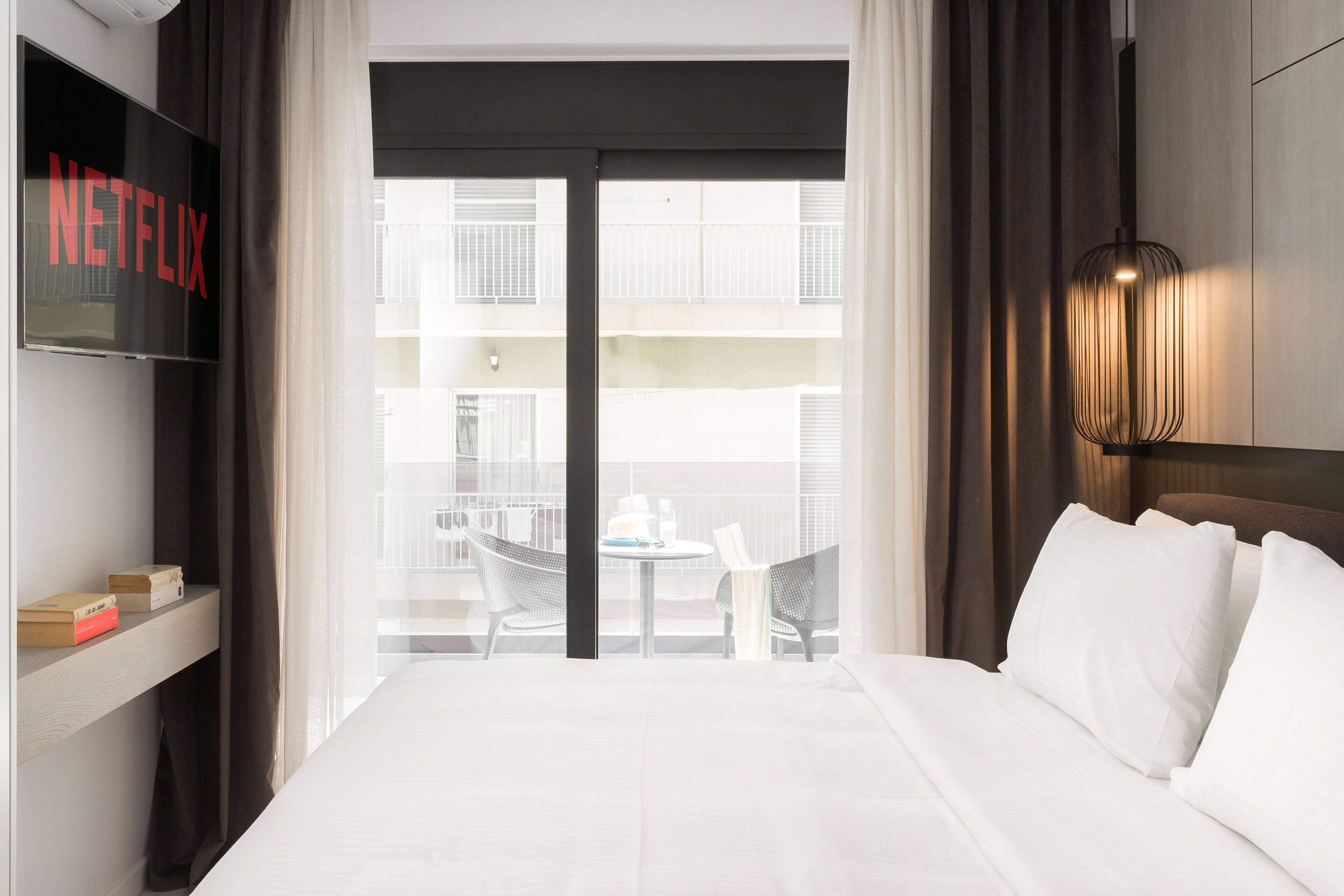A hotel room with a bed, a TV on the wall, a balcony with chairs, and a view of the building.