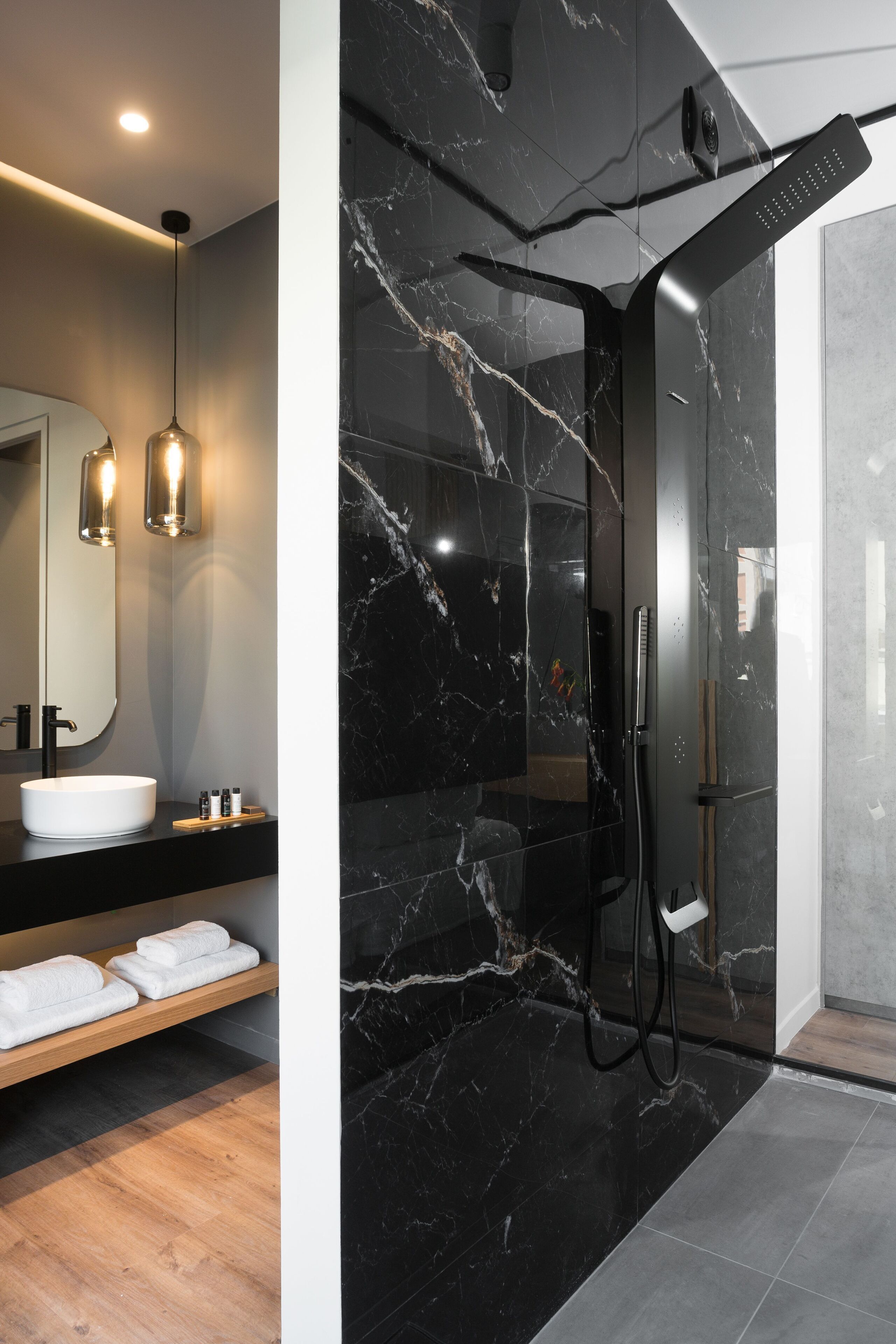 Een moderne badkamer met een douche in zwart marmer, een witte wastafel en een houten plank met handdoeken.
