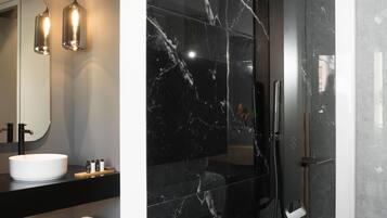 Een moderne badkamer met een douche in zwart marmer, een witte wastafel en een houten plank met handdoeken.
