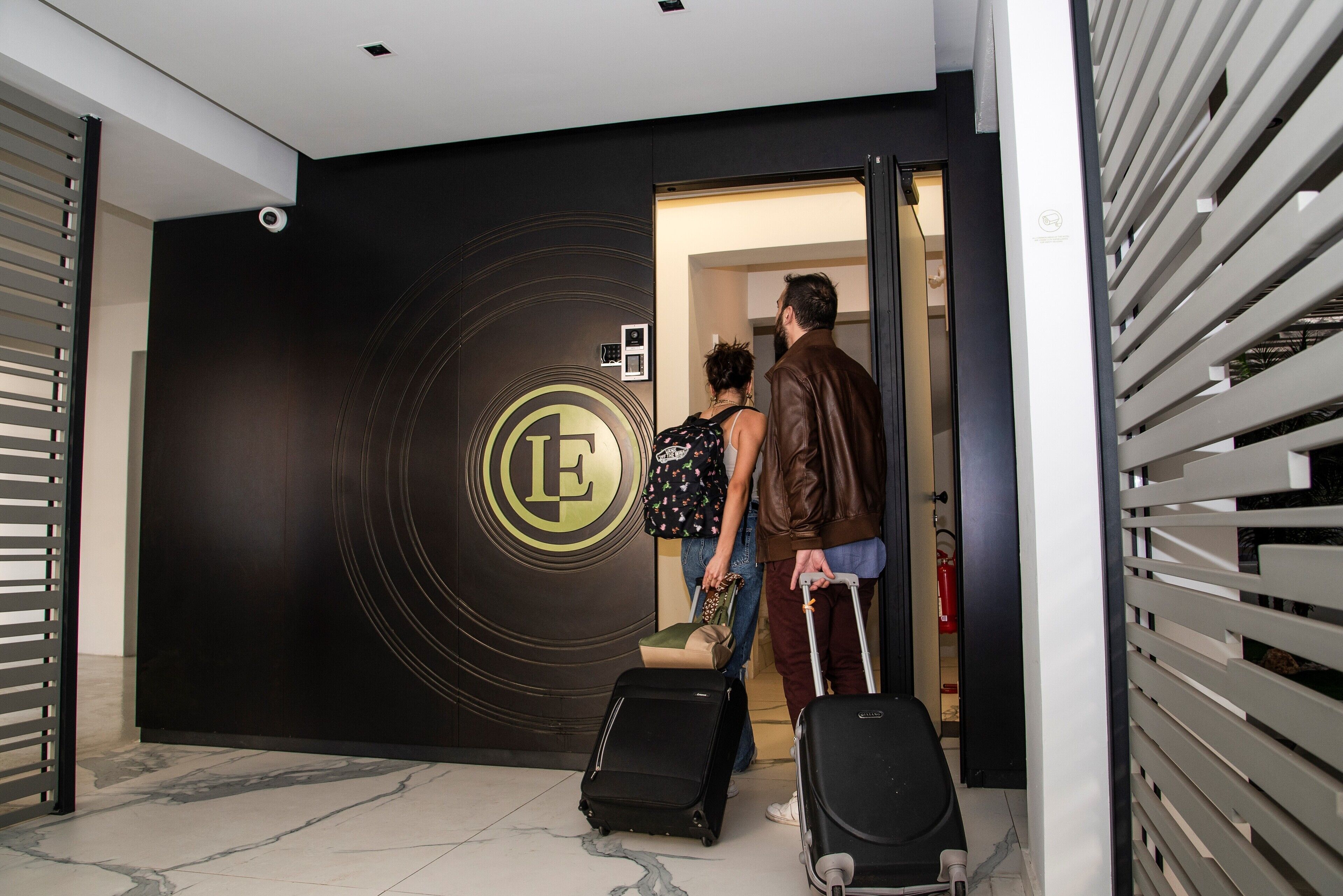 A modern hotel entrance with a large logo on the wall, a security camera, and a fire extinguisher.