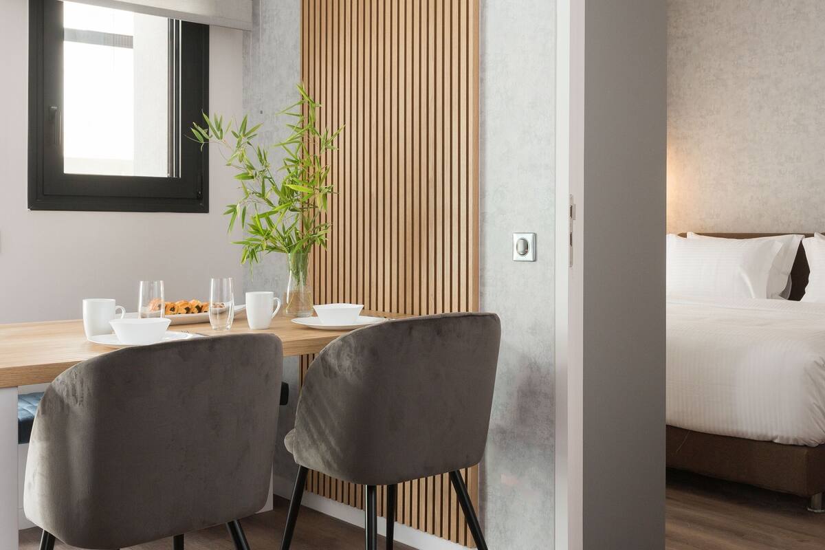 Une chambre d’hôtel moderne avec un séparateur en bois, un coin repas comprenant une table et des chaises, et un lit avec du linge de lit blanc.