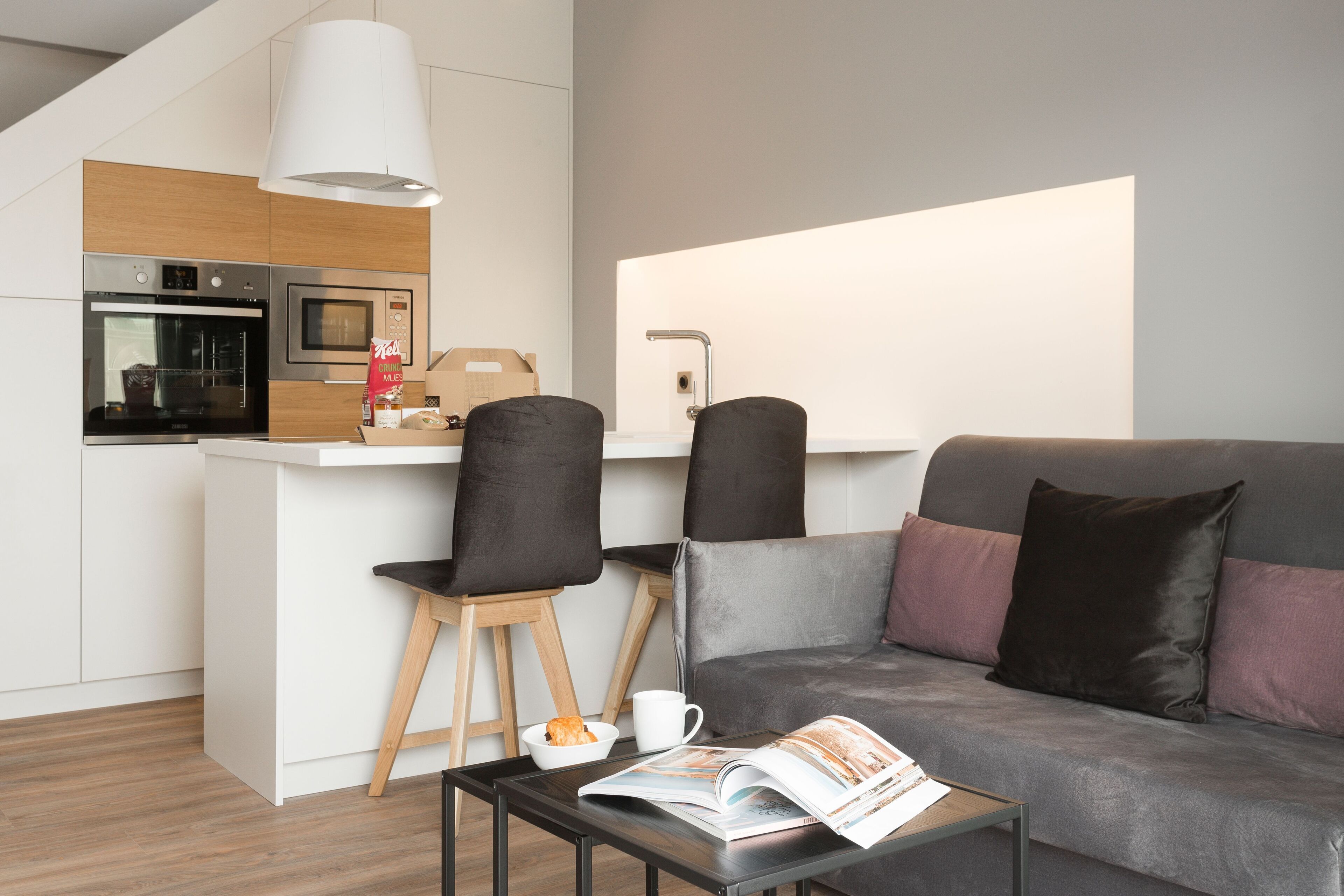 A modern kitchen with a built-in oven and microwave, a dining area with two chairs, a sofa with cushions, and a magazine on a table.